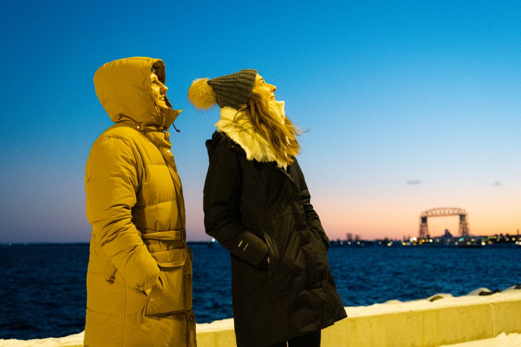 Winter home page 15 Two women dressed for winter by lake superior at dusk with the aerial lift bridge in the background