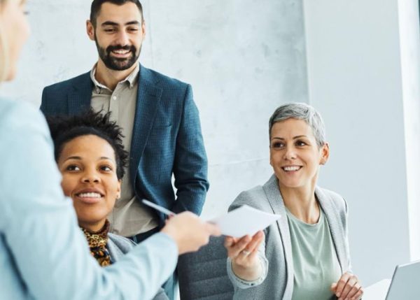 Three people in a meeting room; one hands a document to another, who sits at a table with a laptop.